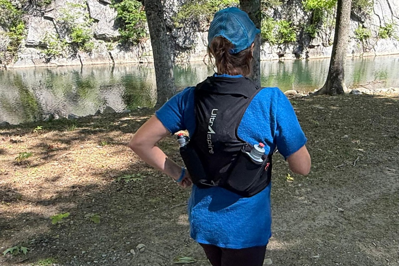Rear view of a woman wearing a black hydration vest running along a forested trail.