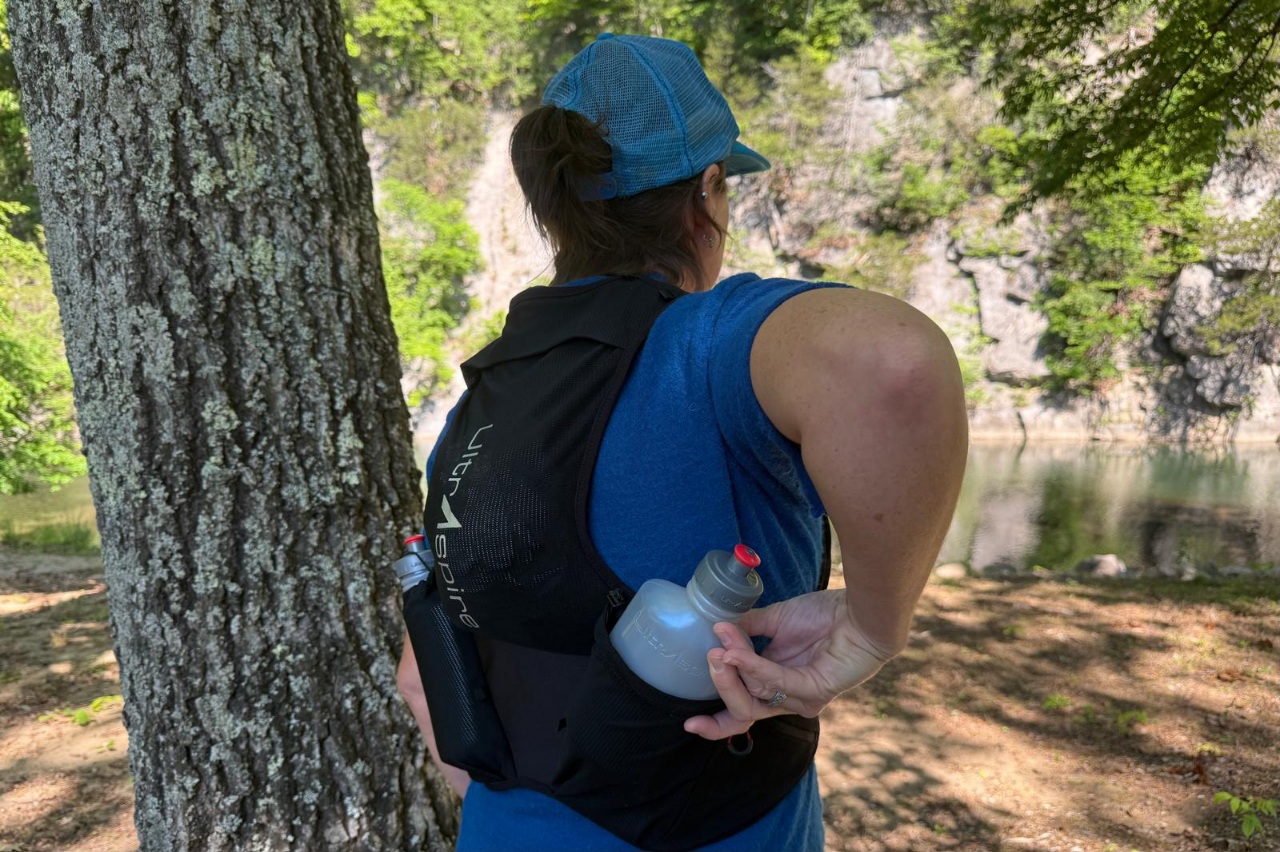 Woman reaching behind her back to grab a water bottle out of a black running vest.