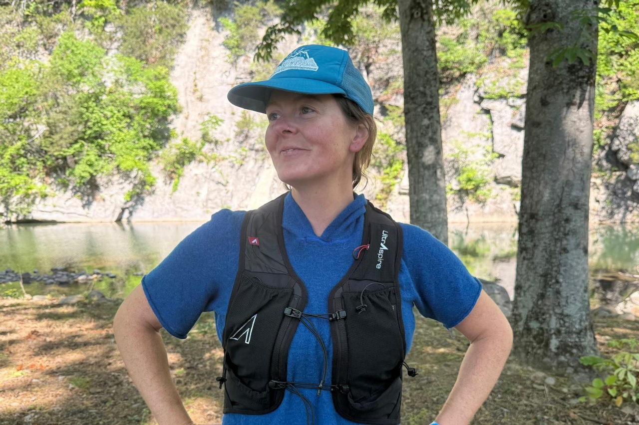 Close-up image of a woman standing on a trail in the forest wearing a black running vest.