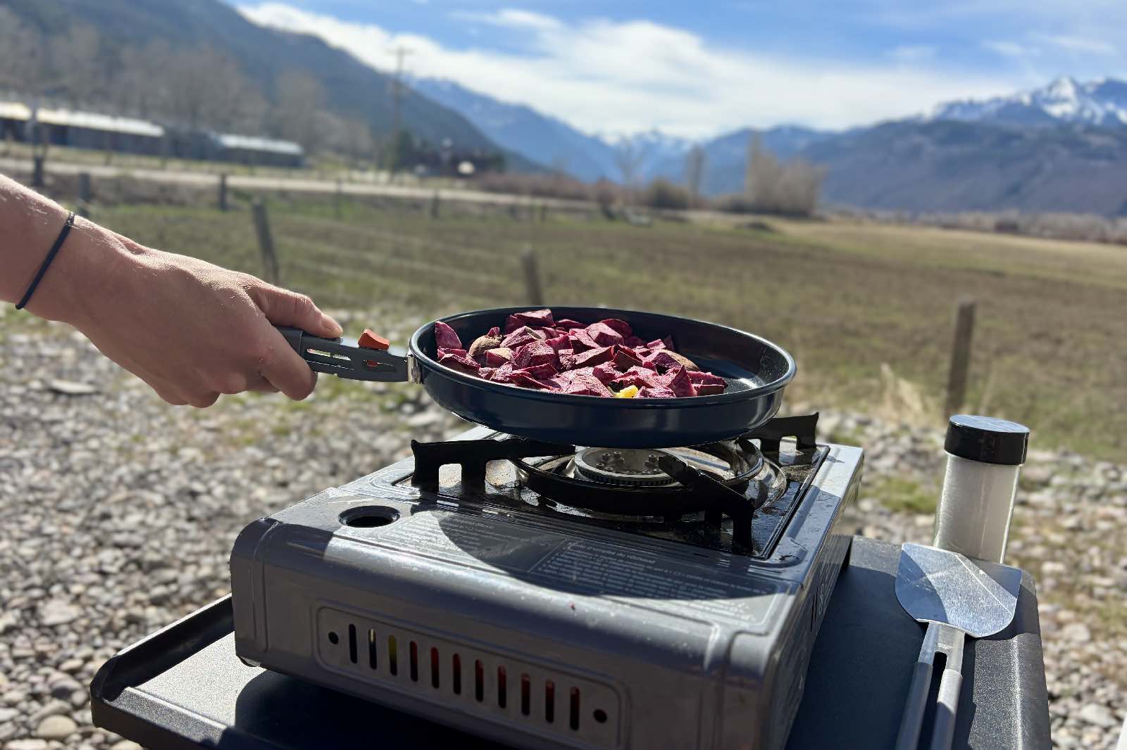 someone holds a frying pan with potatoes over a camp stove