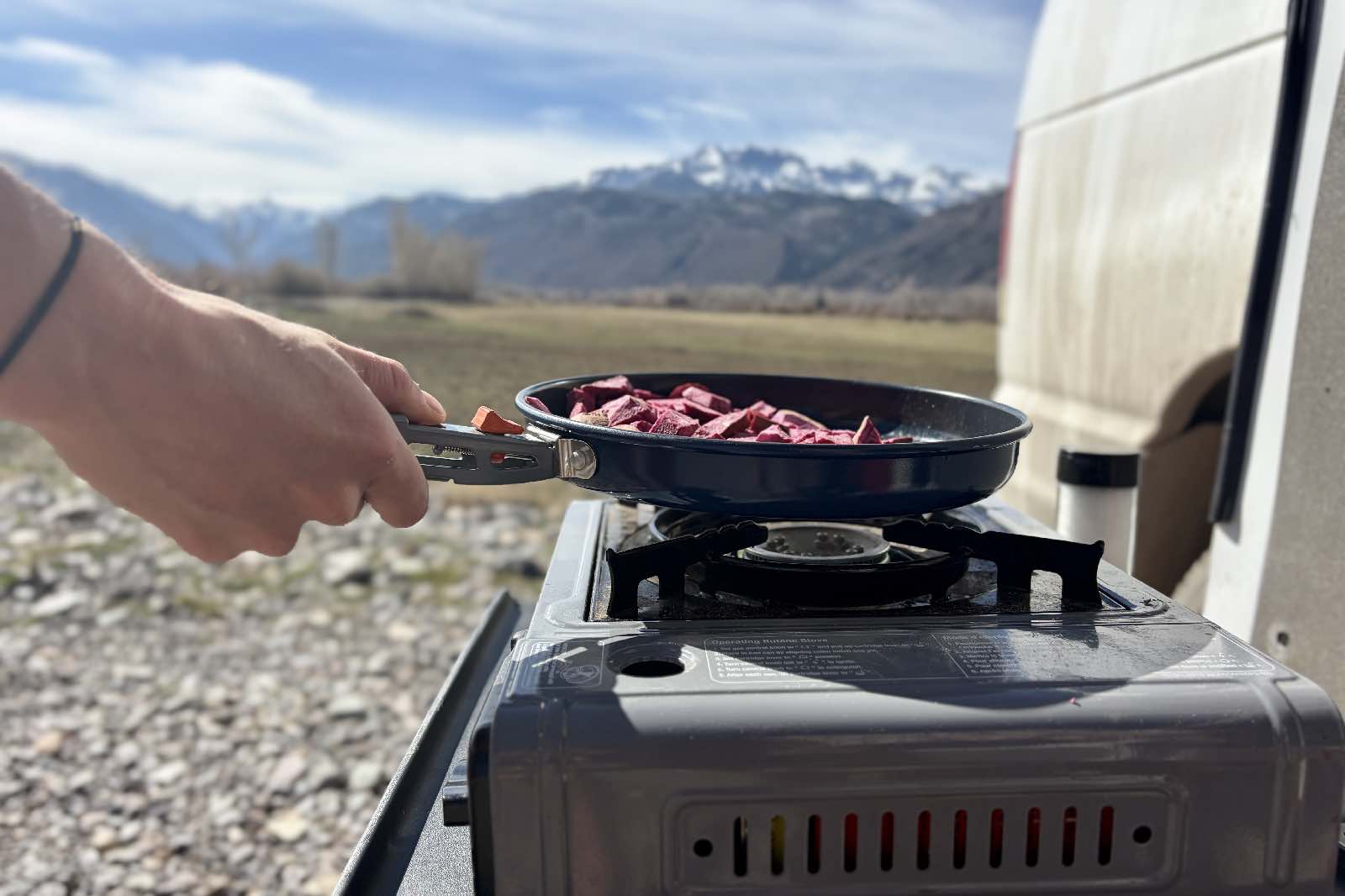 someone holds a frying pan with potatoes over a camp stove