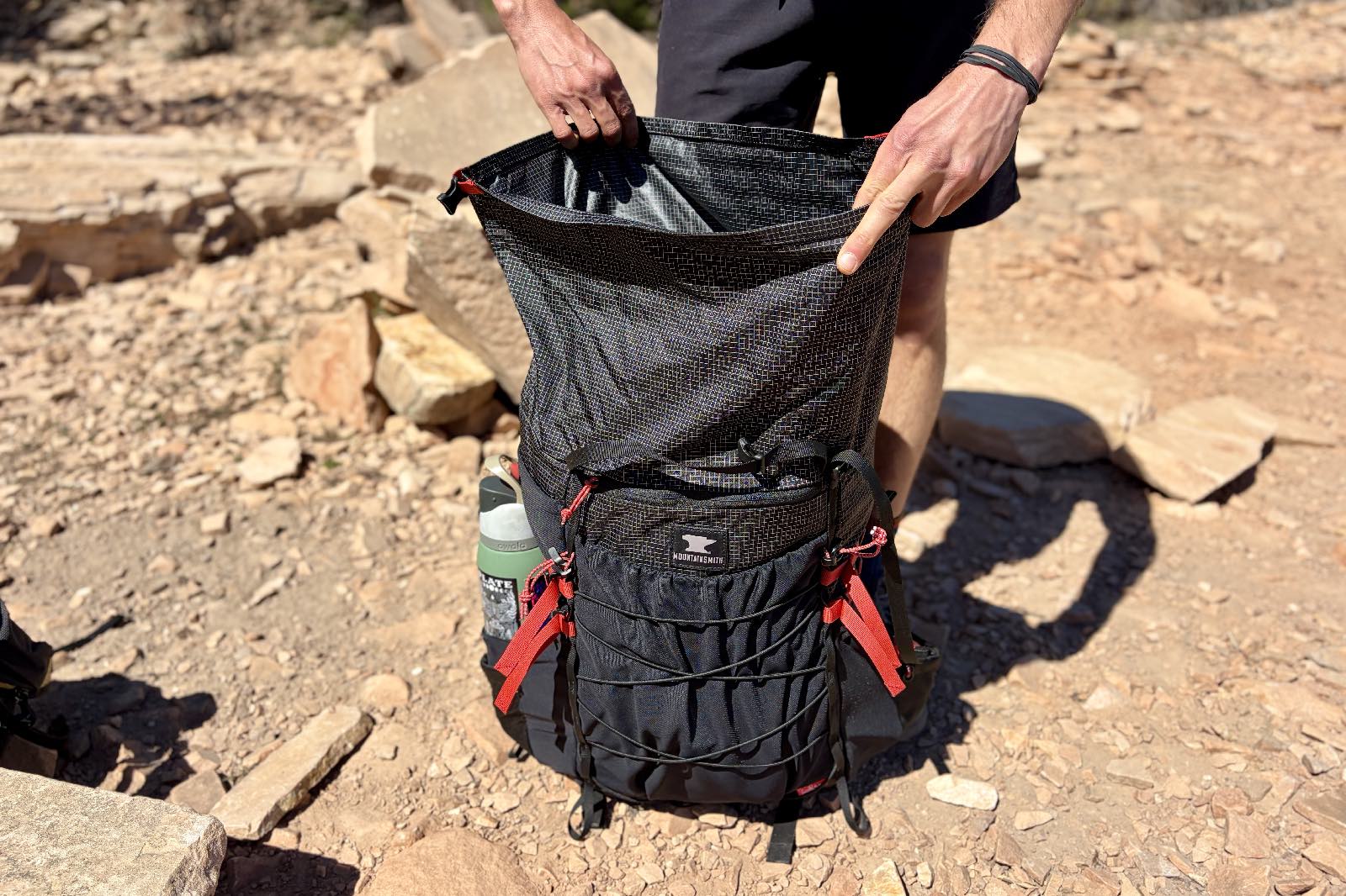 A hiker stands in a trail and looks into the top of a backpack with a roll top enclosure.