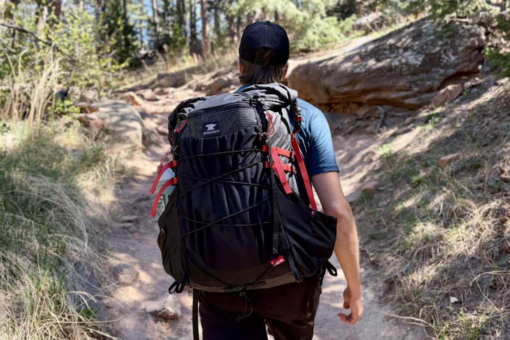 A hiker hikes away from the camera wearing a black backpack with red straps.