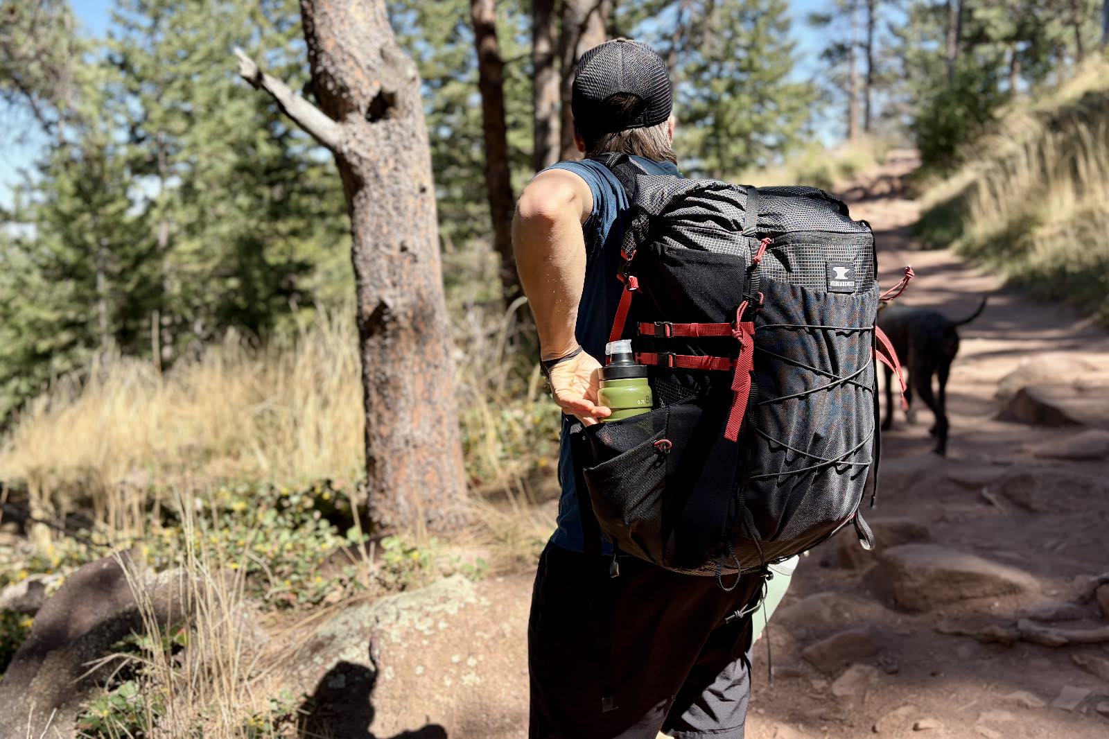 A hiker stands in a trail wearing a black backpack and reaches for his water bottle in the side pocket.