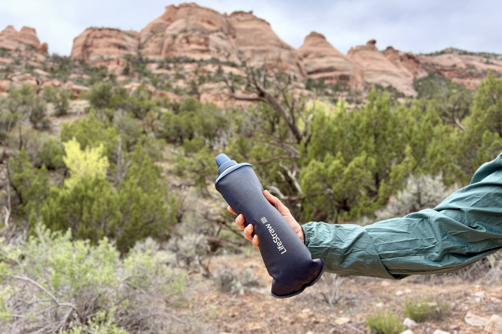 An arm holds a soft flask water bottle in front of a desert background.