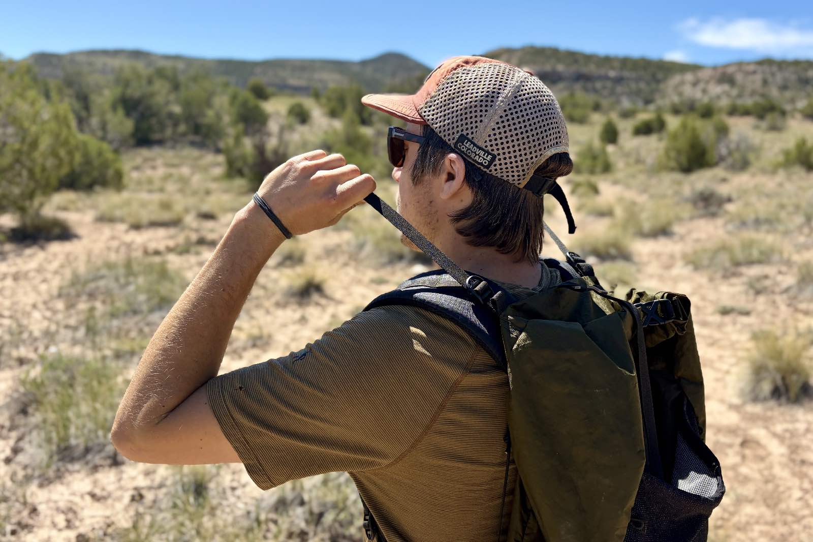 A hiker stands in a desert and pulls the load lifter straps of his green backpack.
