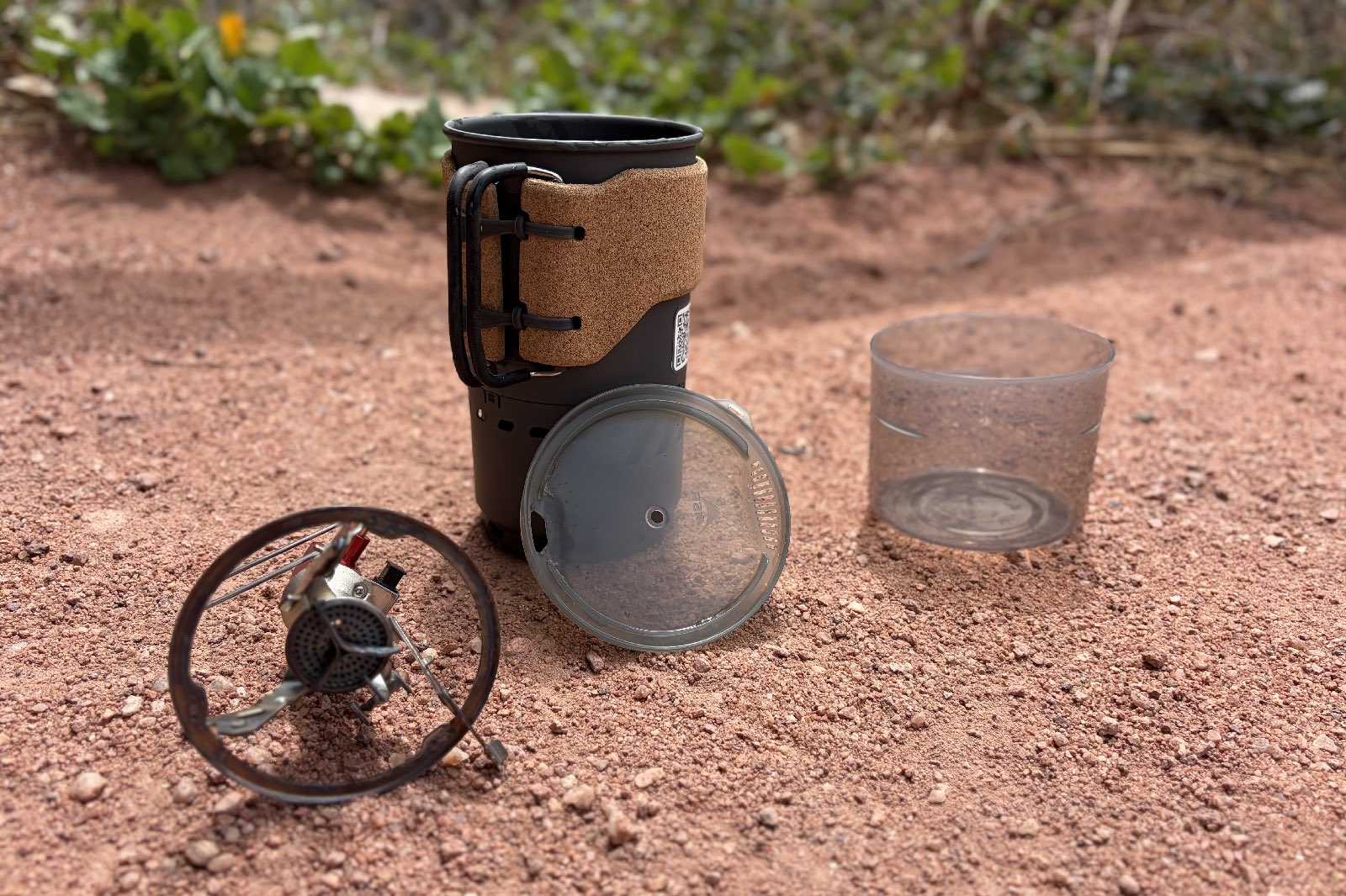 The components of a backpacking stove laid out on red clay ground.