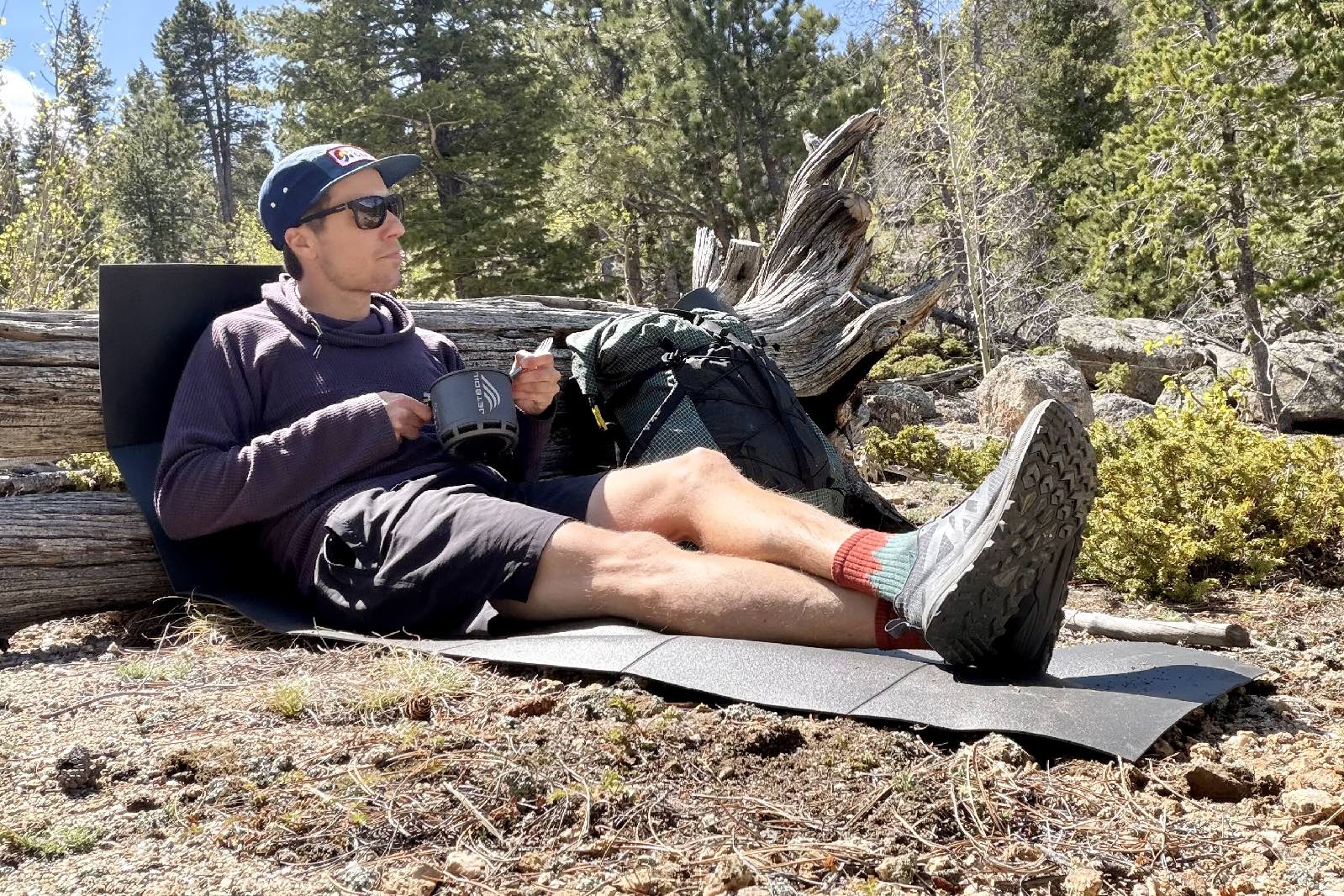 a hiker sits on the ground and leans up against a log with a foam pad underneath him