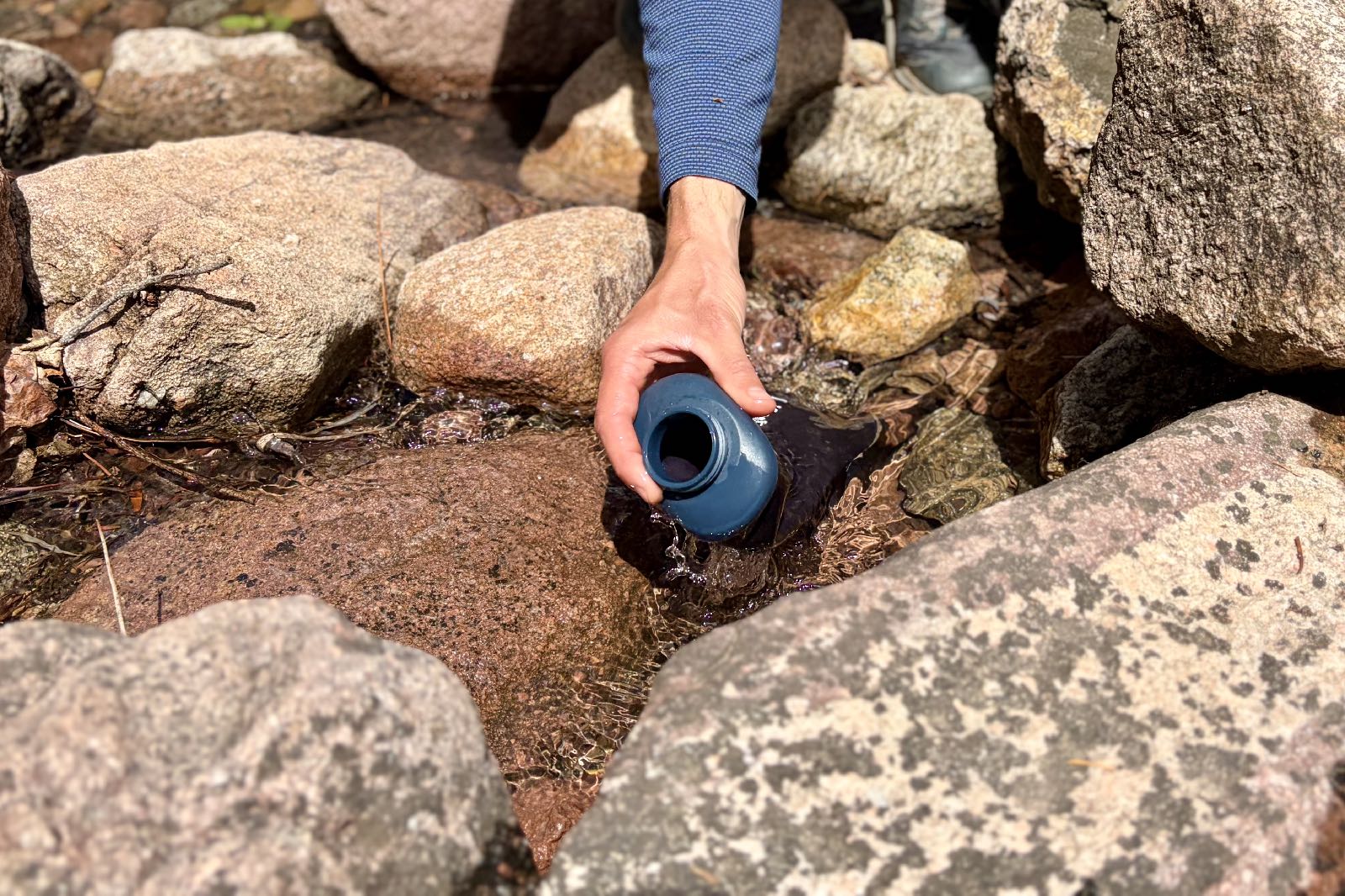 a close up of a hiker scooping water into a soft flask water bottle from a puddle