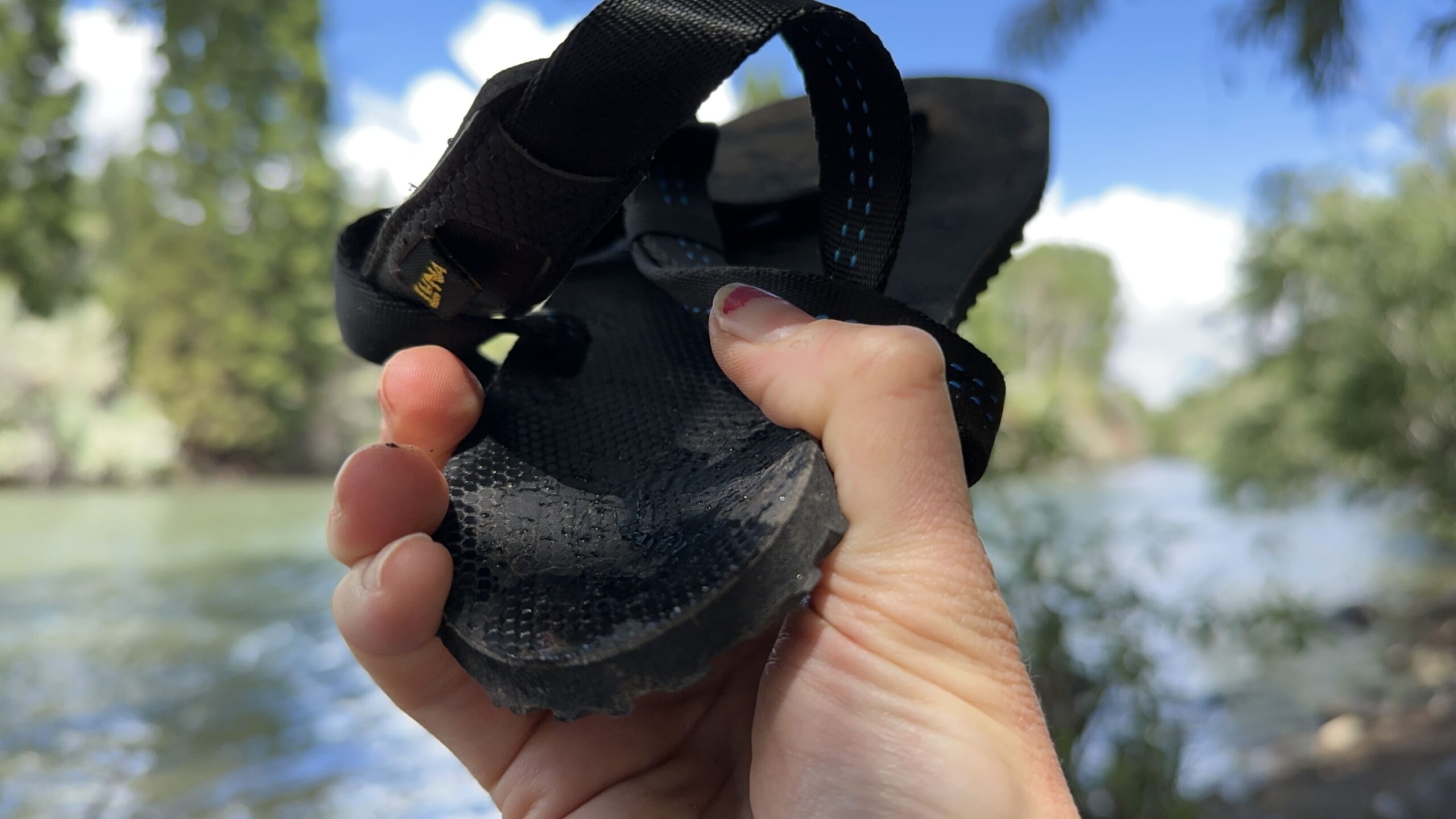 A woman holds a black sandal in front of the camera with a river in the background.