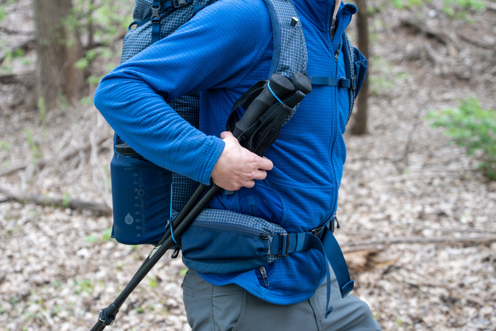 Un hombre sostiene bastones de trekking atados a una mochila azul que lleva puesta.