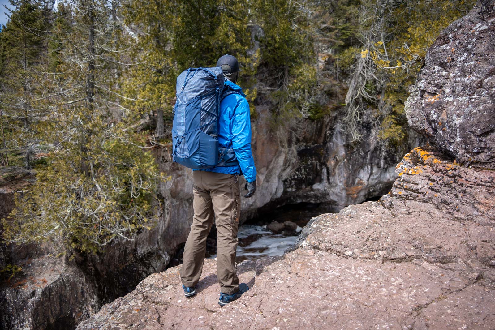 A man wearing a blue backpack stands in front of a river gorge