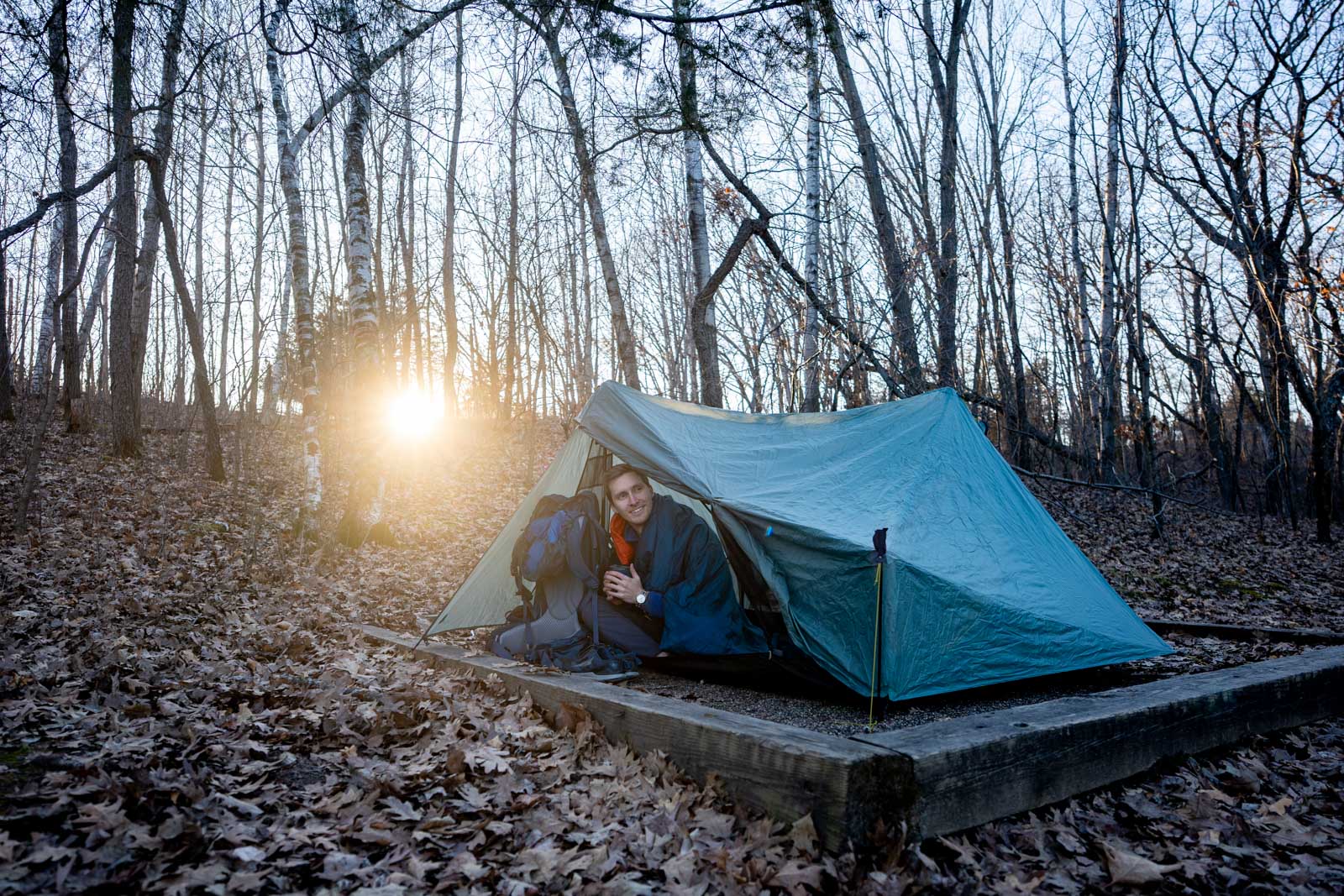 A man holds a mug and sits in a tent vestibule with the sun filtering through the forest behind him