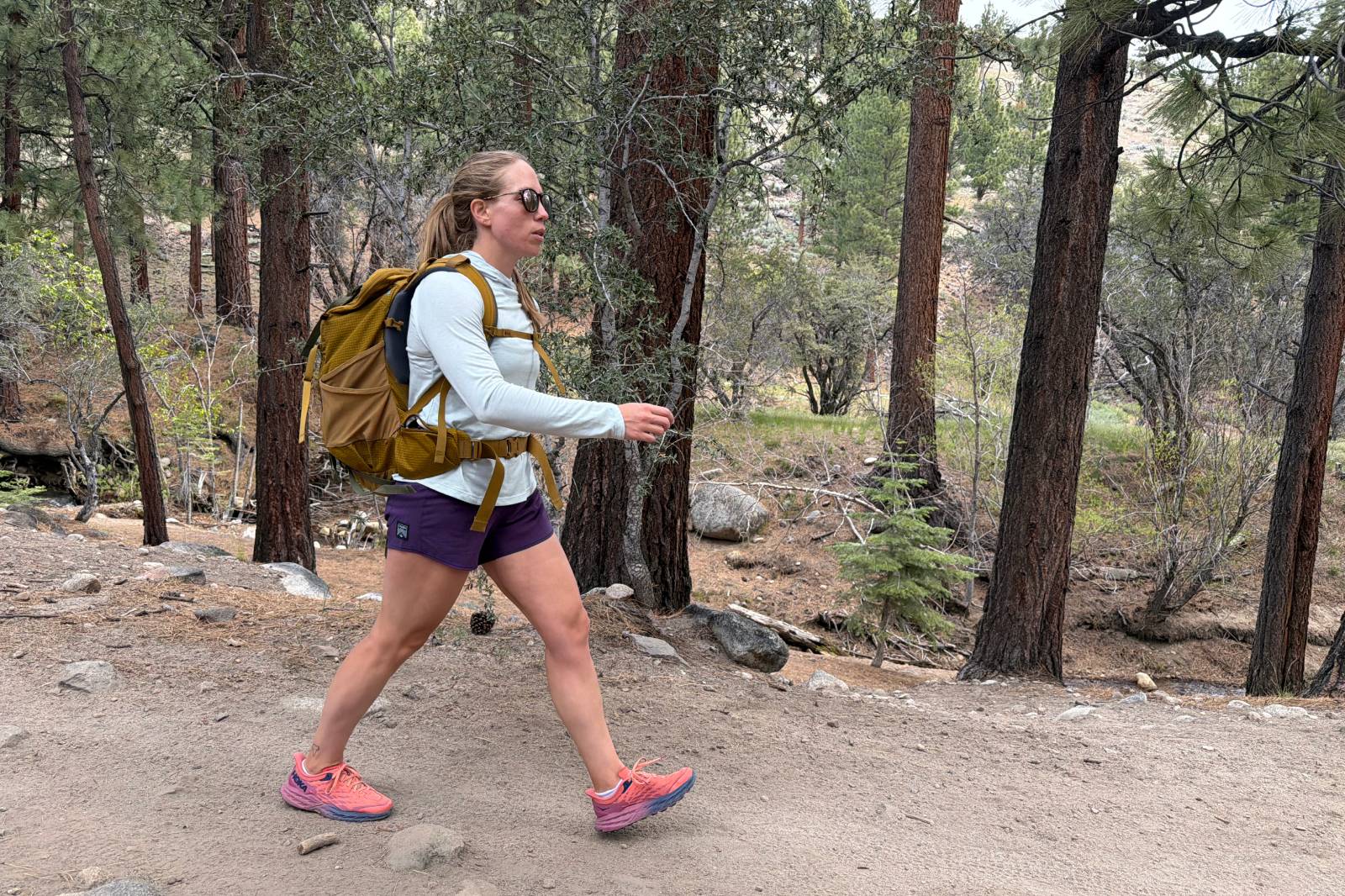 A woman wearing a pair of purple hiking shorts walks along a forested trail.