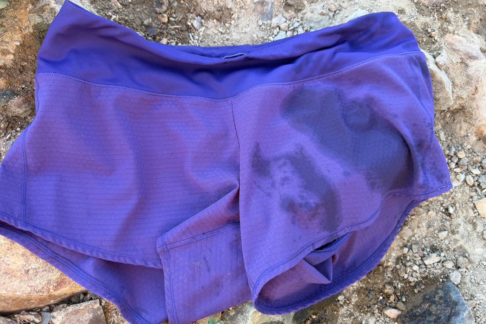 A pair of purple running shorts that's wet on one side lies on a rocky trail.