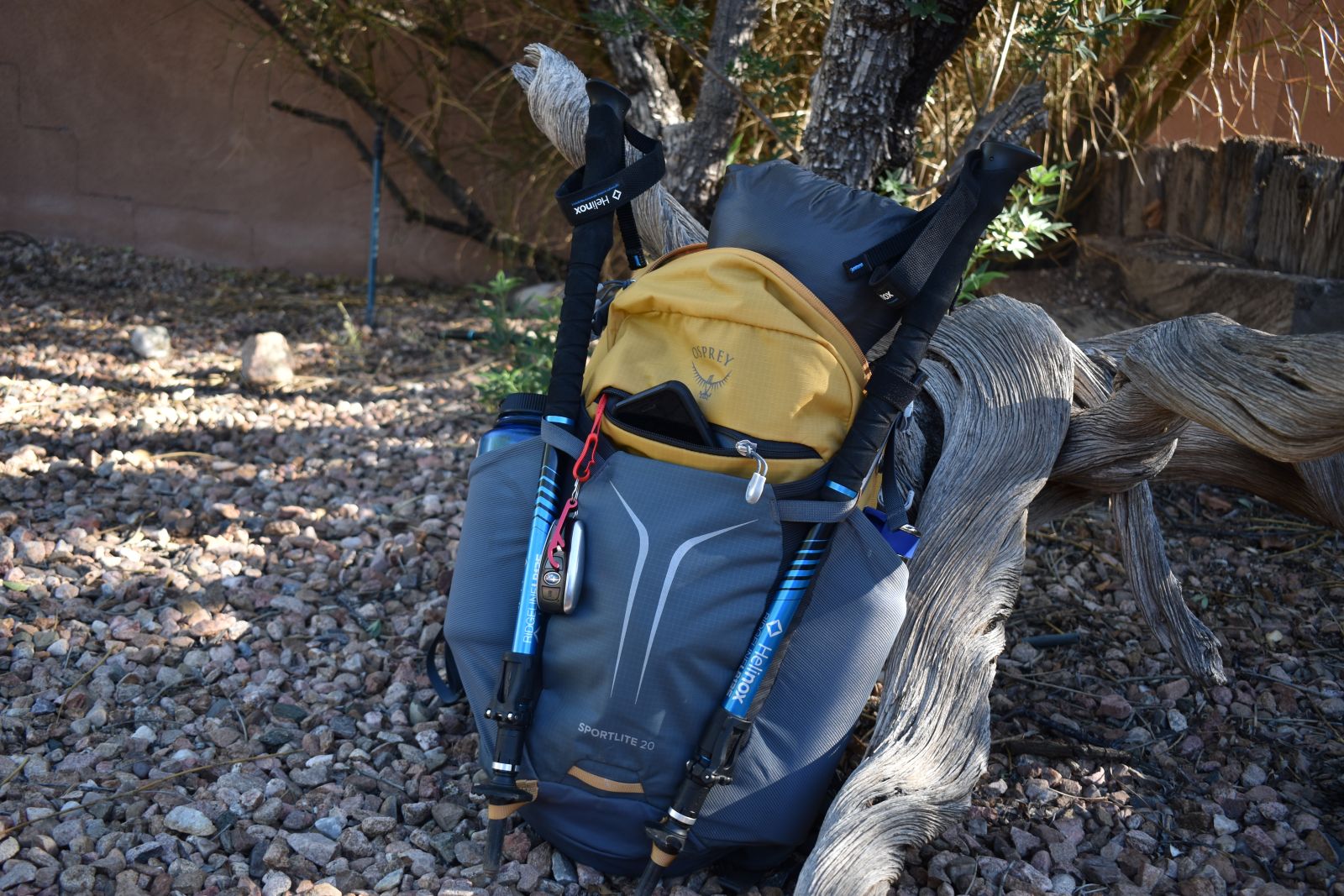 The Osprey Sportlite 20 lays against driftwood with layers, trekking poles, water bottles, and personal items stick out of their pockets and bungees.