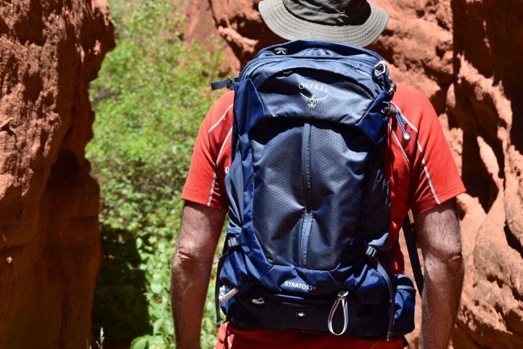 Close up of a man in a red hiking shirt wearing a blue Osprey Stratos daypack in a redrock slot canyon