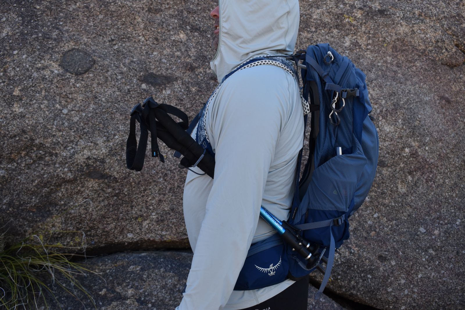 Profile view of a male hiker in a light hoodie wearing the Osprey Stratos. His treking poles are attached on the side, and space is visible between his back and the backpack.