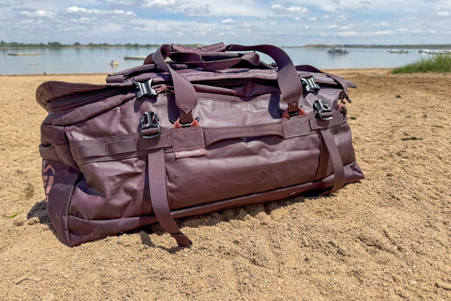 REI Co-op Big Haul 60 duffel sitting on sand near a lake with boats in the background.