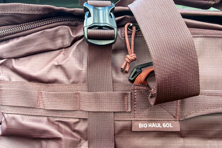 Close-up of the REI Co-op Big Haul 60 showing the compression strap, buckle, zipper pull, webbing, and product label.