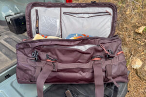 REI Co-op Big Haul 60 duffel opened in a truck bed, showing the wide main compartment, interior mesh pockets, exterior pocket, and compression straps.
