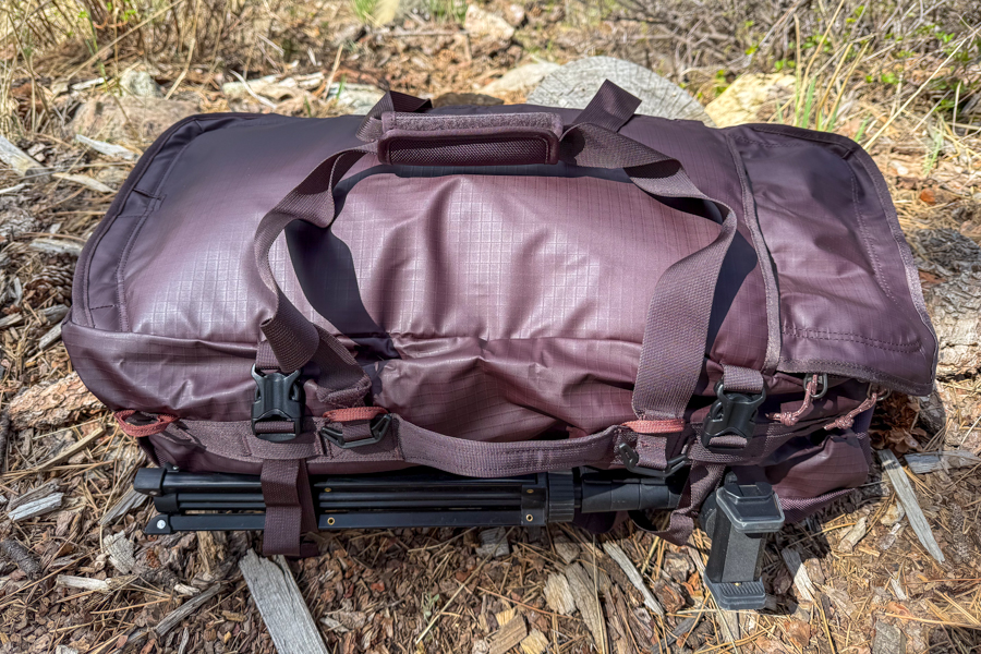 REI Co-op Big Haul 60 strapped to a folded camp table on the ground, showing compression straps, buckles, and tote handles.