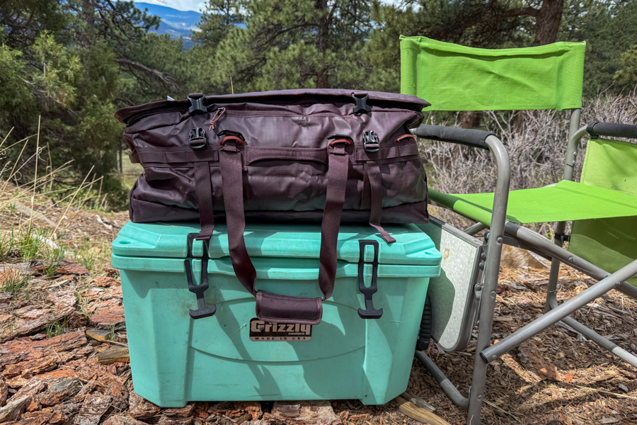 REI Co-op Big Haul 60 duffel sitting on a green cooler next to a camp chair at a wooded campsite.