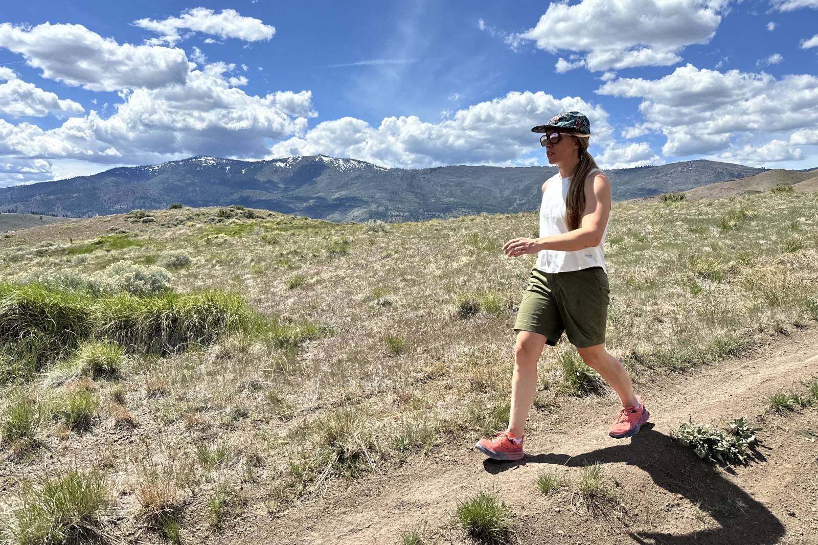 Una mujer que lleva un par de pantalones cortos de senderismo verdes hasta las rodillas camina por un estrecho sendero de tierra con montañas al fondo.