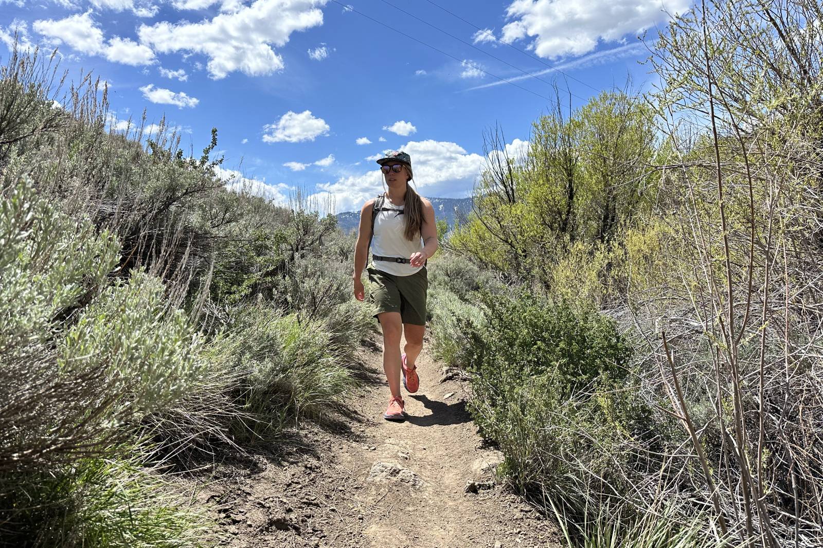 A woman wearing a pair of green knee-length hiking shorts hikes toward the camera on a trail with brush on either side.