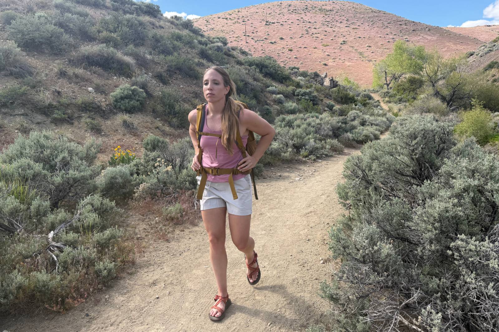 A woman wearing a pair of beige thigh-length hiking shorts walks along a dirt trail with desert shrubs in the background.