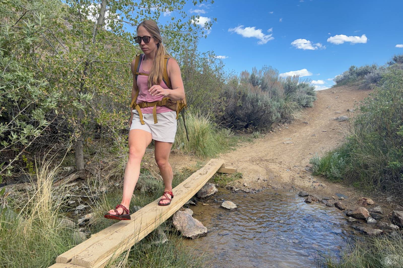 A woman wearing a pair of beige thigh-length hiking shorts walks across a plank to avoid stepping in a creek.