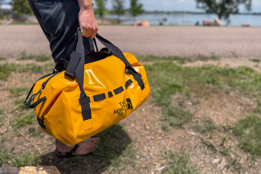 Person carrying the yellow The North Face Base Camp Waterproof Duffel 50L by its tote handles near a lake.