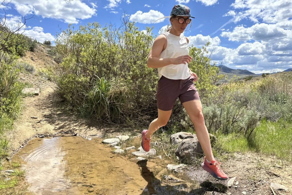 A woman wearing a pair of thigh-length hiking shorts takes a long stride to clear a shallow creek.