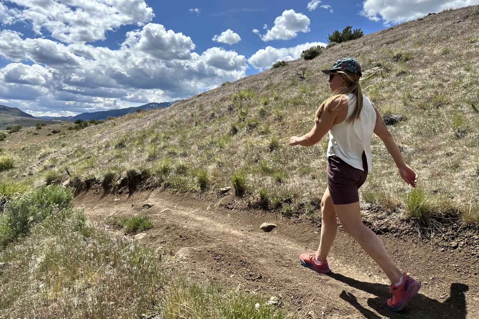 A woman wearing a pair of thigh-length hiking shorts walks along a dirt trail on a sunny day.