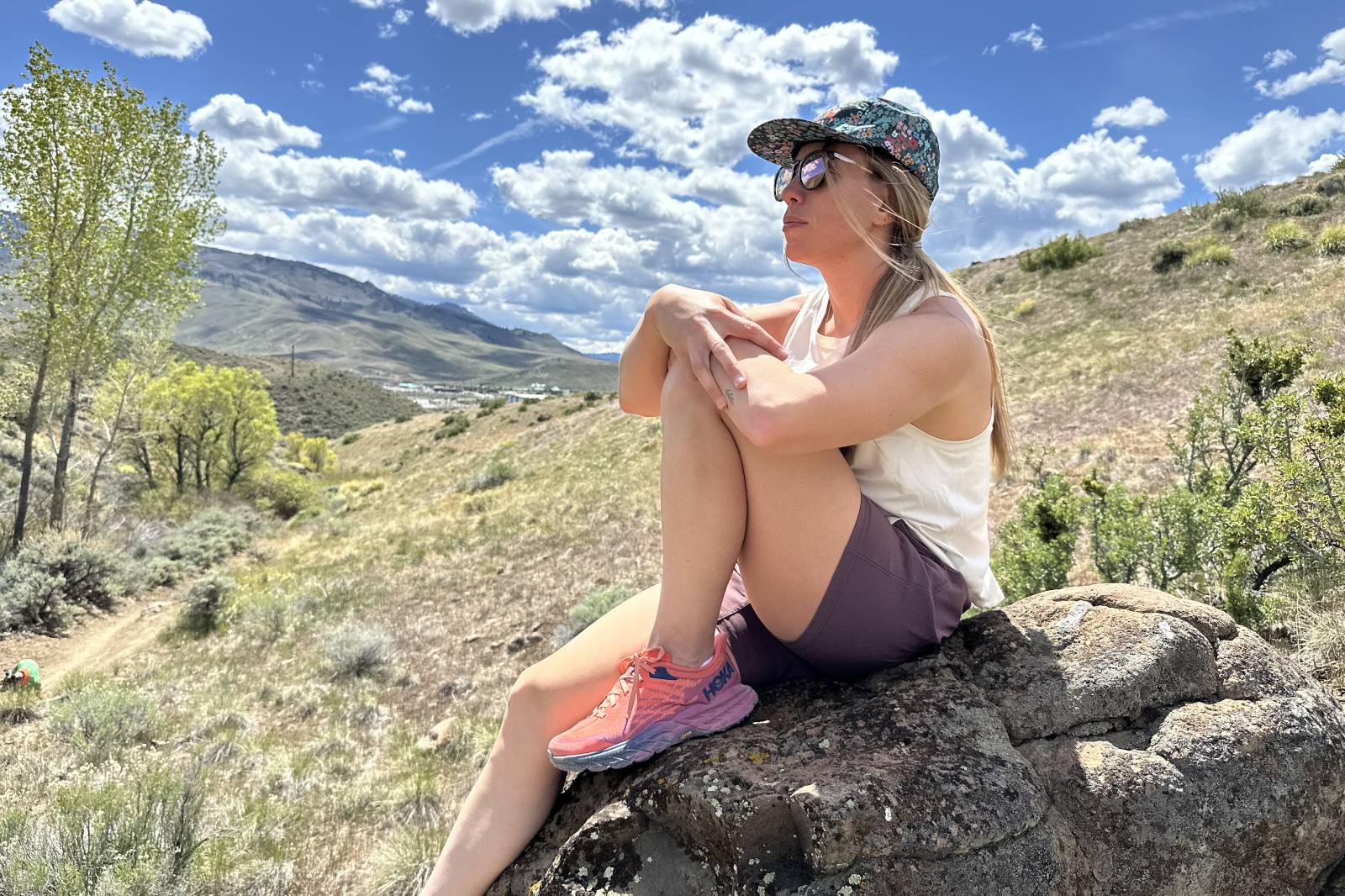 A woman wearing a pair of hiking shorts sits on a rock hugging one knee.