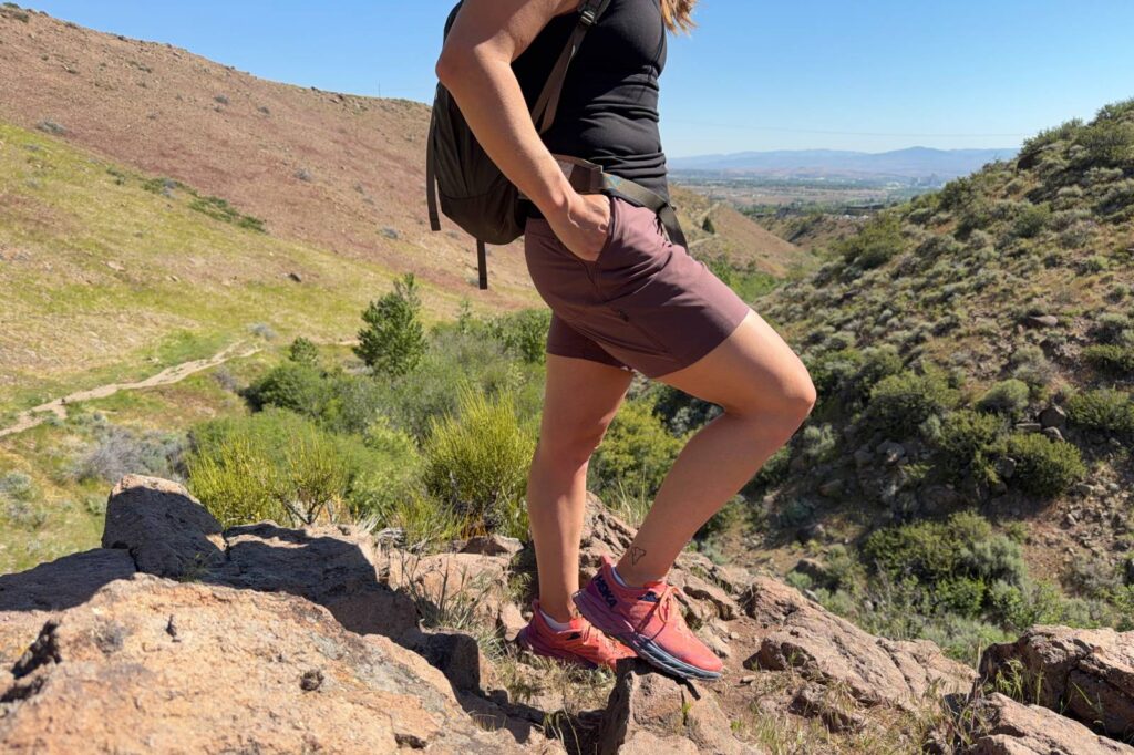 Close-up image of a woman in hiking shorts standing on a rocky outcropping with a view of the trail and forest below.