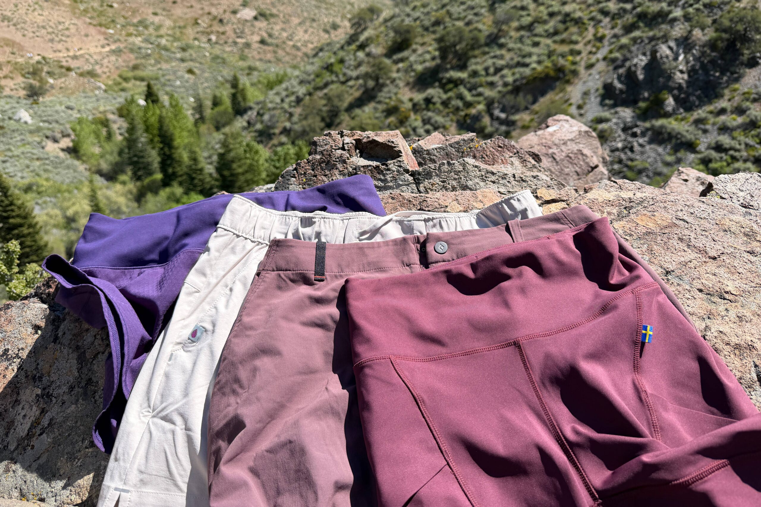 Close-up image of the waistbands of four pairs of hiking shorts laid out on a rock with a blurry forest in the background.