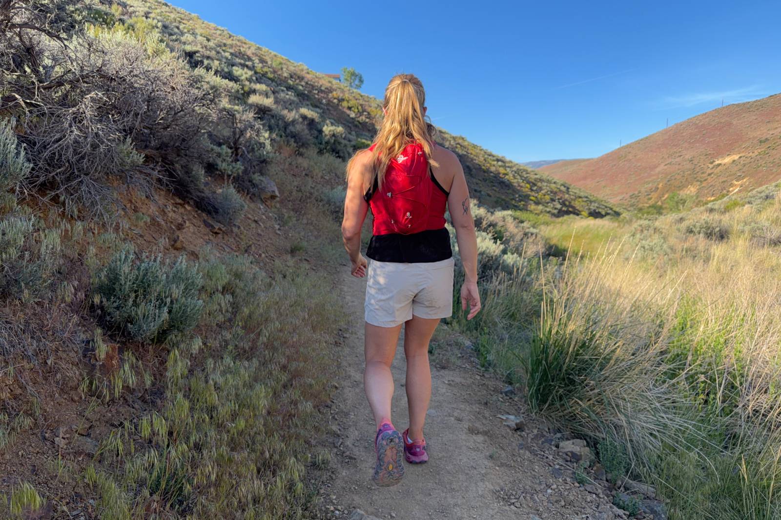 A woman wearing beige hiking shorts walks away from the camera on a dirt trail with brush on either side.