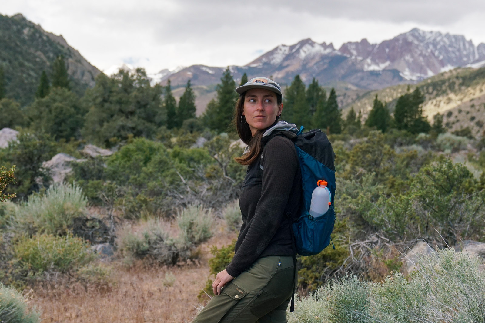 A person wearing a backpack stands on a trail with mountains and bushes in the background.