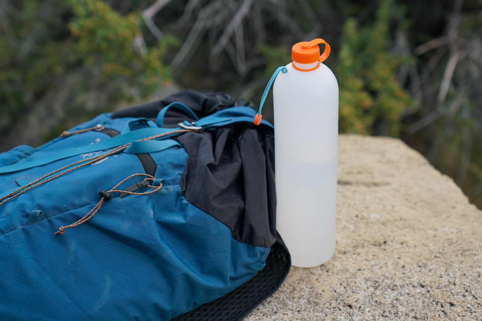 A close up of a water bottle sitting on a rock next to a backpack.