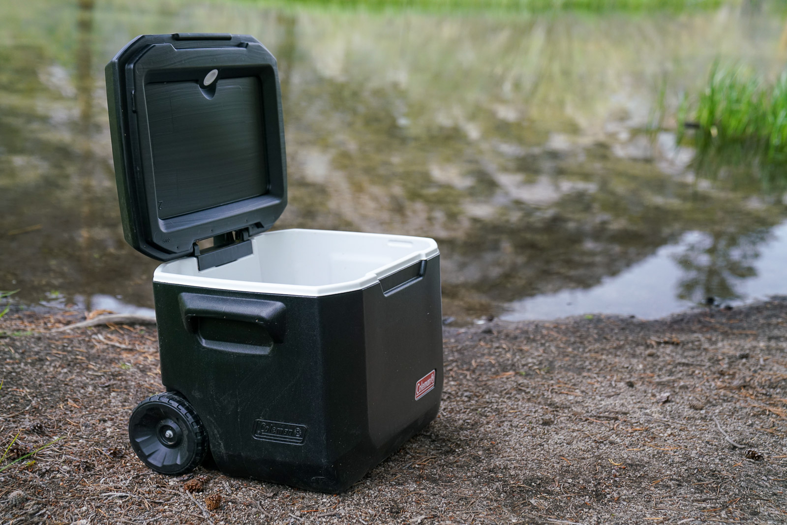A close up of a cooler with its lid open sitting on the ground near a lake.