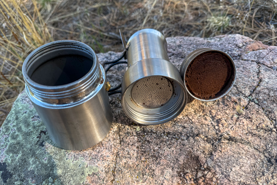A coffee maker disassembled after brewing, showing the used coffee puck, lower chamber, and filter section on a rock.