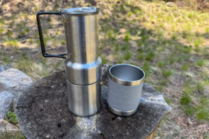 A coffee maker disassembled to show the nesting mug and stainless steel brewer components side by side on a tree stump.