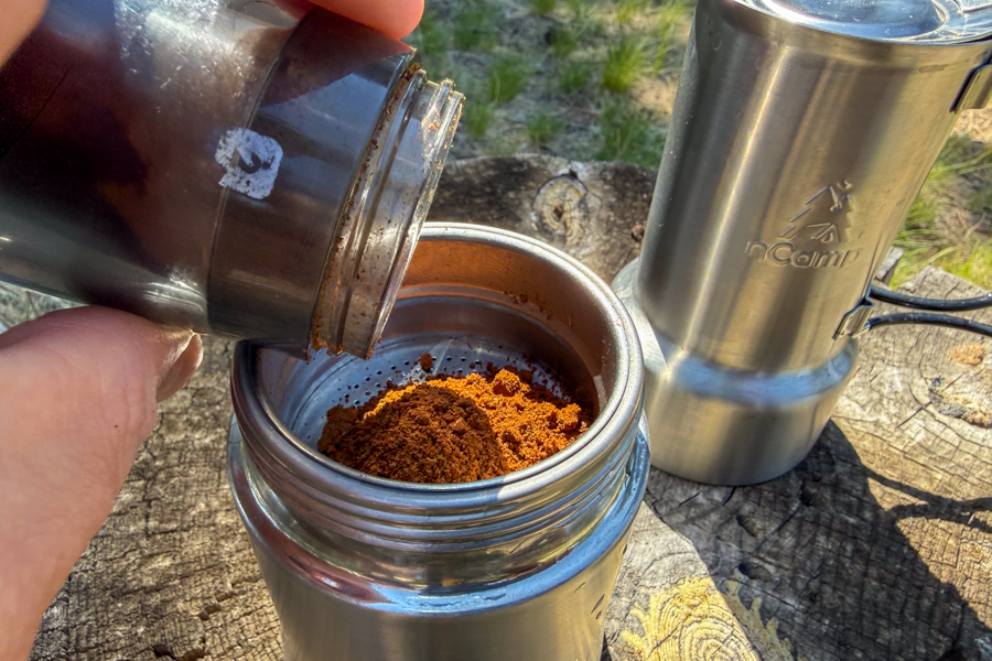 Ground espresso being poured into a filter basket beside a stainless steel coffee maker body.