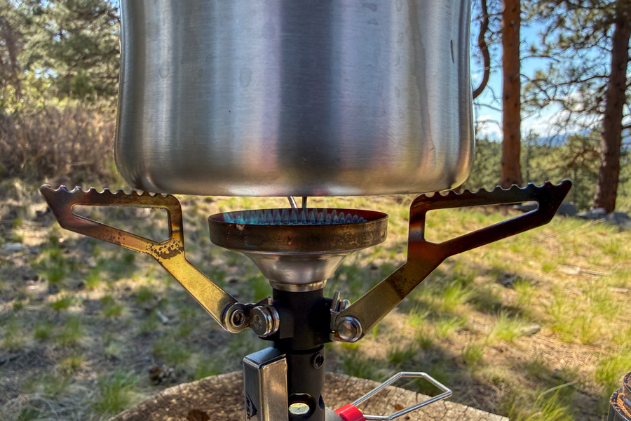 A coffee maker brewing on a small canister stove, with the narrow stainless steel base centered over the flame.