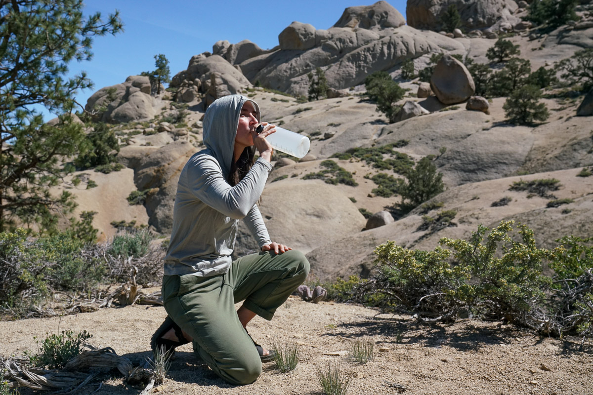 A person kneels and drinks from a water bottle with a rocky landscape in the background.