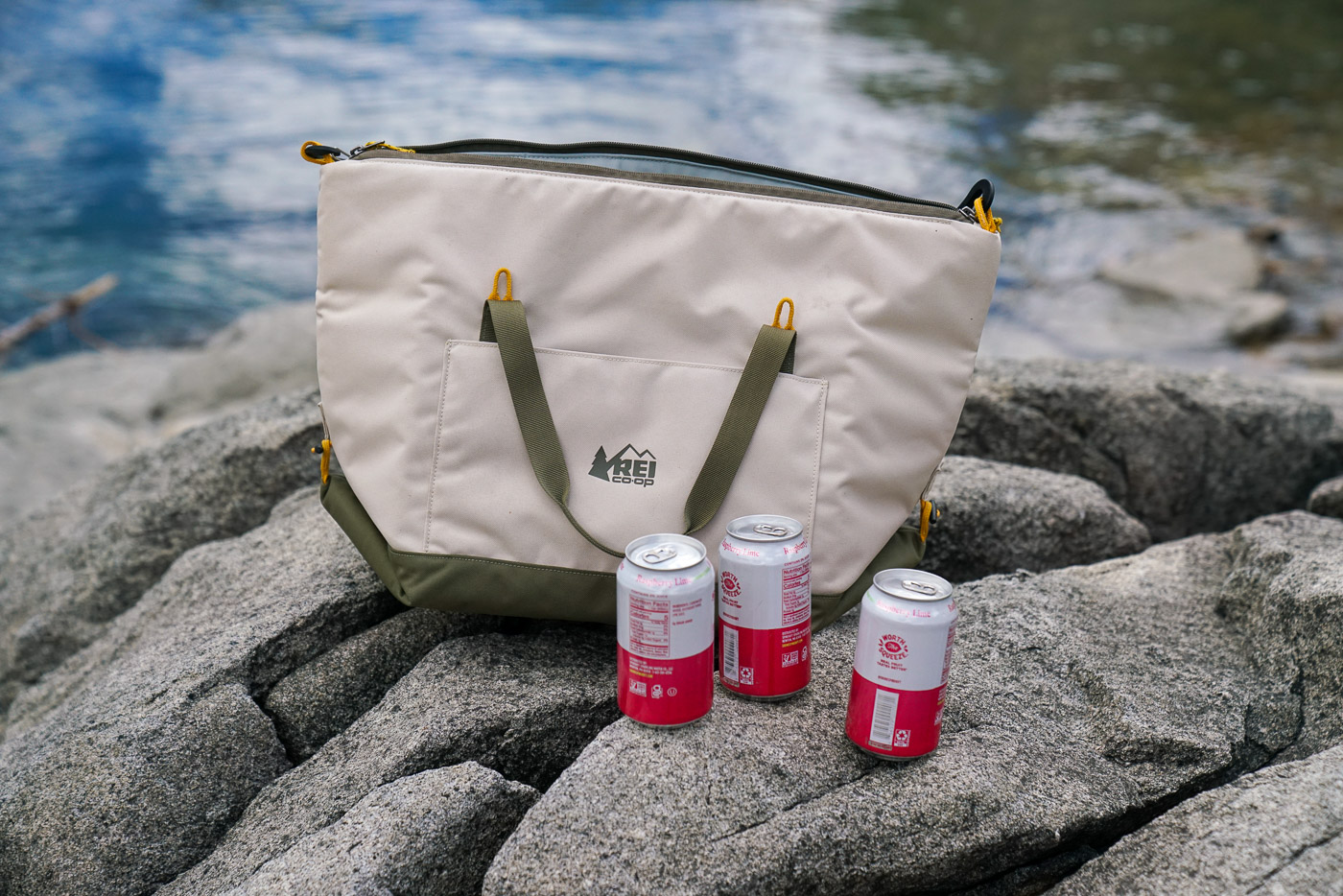 A close up of three canned drinks and a soft-sided cooler sitting near the water.