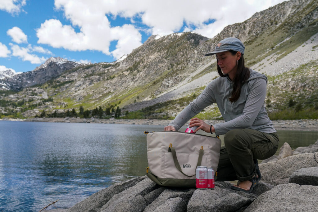 A person places a can inside a soft-sided cooler. There are mountains and a lake in the background.