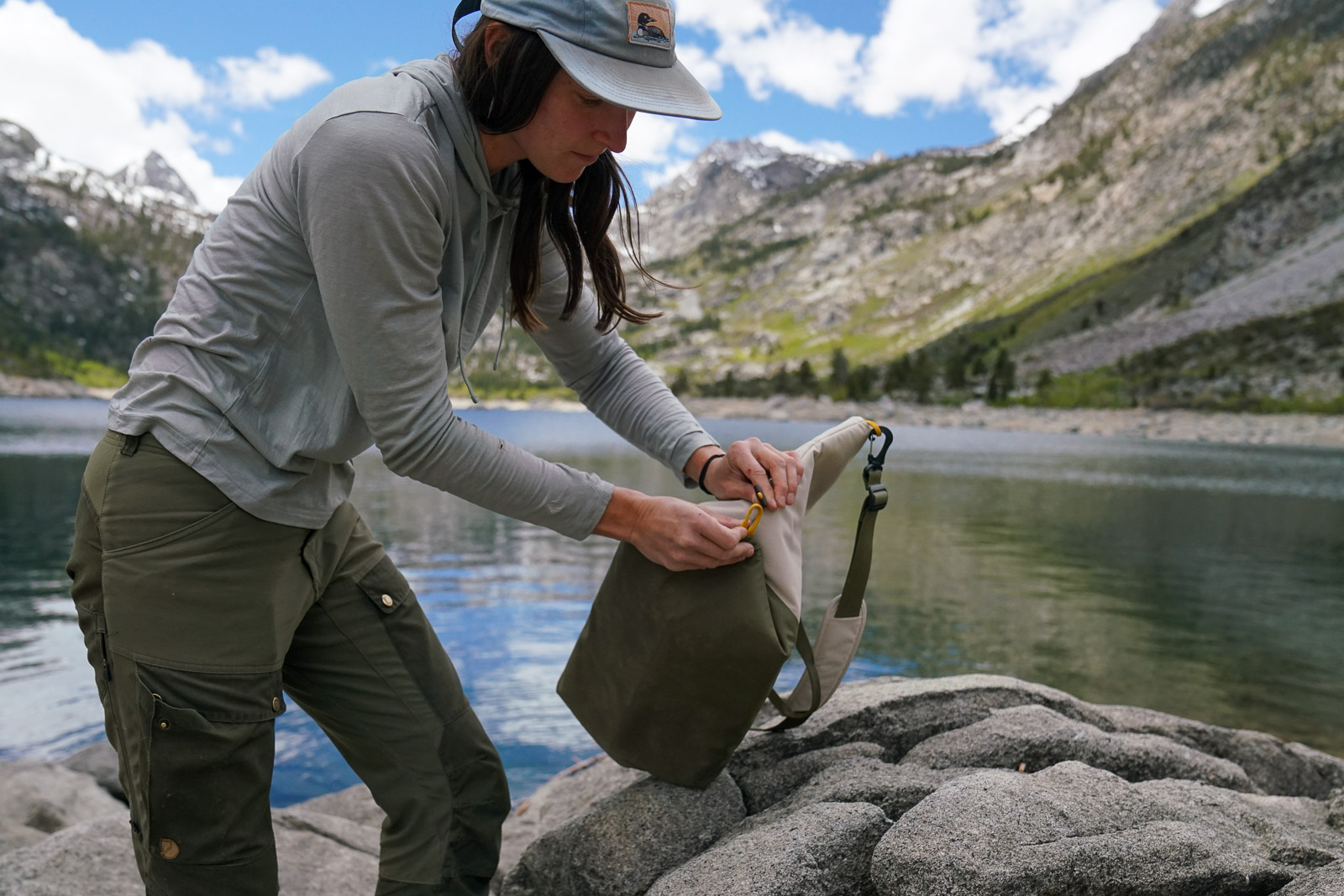 A person hooks a corner of a soft-sided cooler around a toggle. There is a lake and mountains visible in the background.