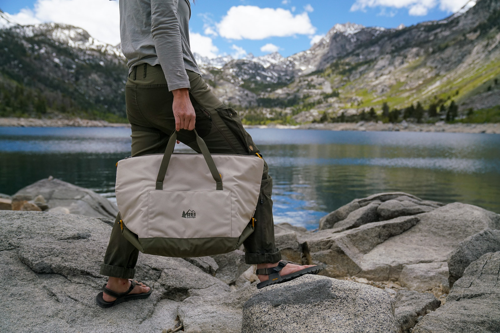 A close up of a person holding the handles of a soft-sided cooler. They walk on a rocky shore next to a mountainside lake.